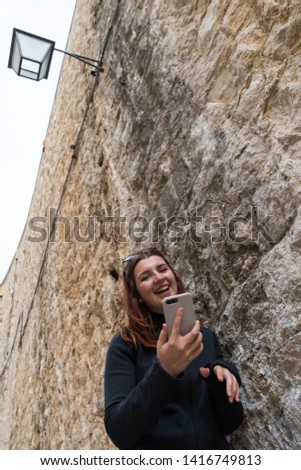 Beautiful young tourist woman visiting city sightseeing on stone wall, holding smartphone taking selfies photos, networking outdoors. Female using technology, fun travel leisure recreation lifestyle.