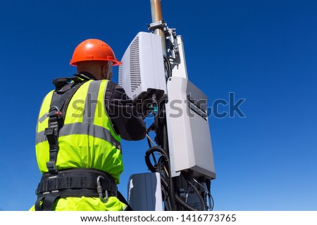 Telecommunication engineer in helmet and uniform installs telecomunication equipment in his hand and antennas of GSM DCS UMTS LTE bands, outdoor radio units, optic fibers, power cables are installed Royalty-Free Stock Photo #1416773765