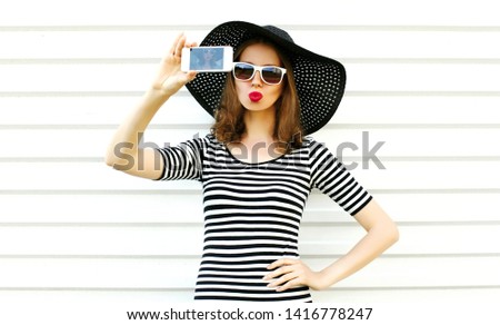 Beautiful woman taking selfie picture by phone blowing red lips sending sweet air kiss on white wall background, female model wearing black summer straw hat