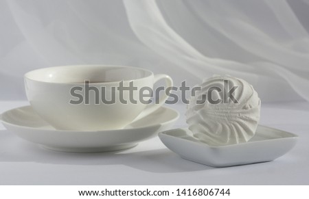 One marshmallow on a squre small plate and white cup of black tea on a plate on a white with veil on the background. Natural daylight.