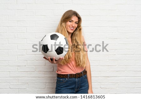 Young blonde woman over white brick wall holding a soccer ball