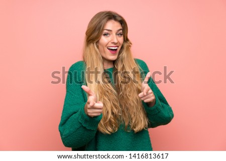 Young blonde woman with green sweater over pink wall points finger at you