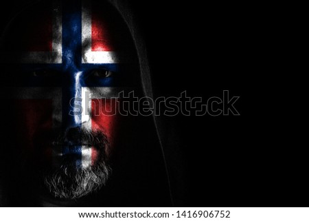 A brutal man with a gray beard, with the flag of Norway on his face, in a hood with sharp shadows on a black background. Copy space