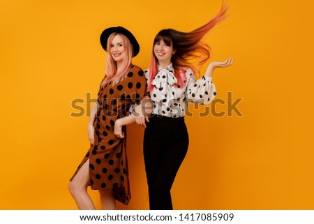 Two friends, pretty girls walking and having fun on yellow background in studio. Happy woman with pink hairs  with her blond friend  having great time.