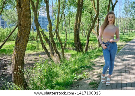 Photograph of a beautiful girl woman on nature in the park with long flowing beautiful hair in a pink sweater and blue jeans on a sunny day is standing right in front of the camera and smiling.