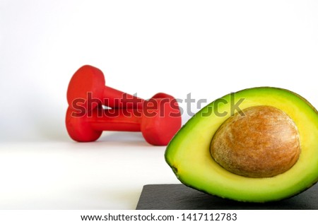 Fresh cut avocado with unfocused red dumbbells on background. Concept of healthy nutritious eating and fitness. 