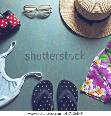 Summer holiday preparation checklist - Straw boater hat with black ribbon, plastic heart shaped glasses, toy camera, flip flop sandal, white camisole, colorful Hawaiian shirt on blue background.