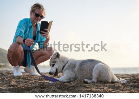 Young pretty blond woman, taking picture on her phone of her small husky puppy. Owner and the light grey dog, playing on the sandy beach at sunset. 
