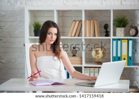 A young brunette girl in a white dress is sitting in the office at the table. Woman works at the laptop