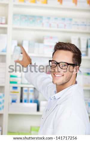 Portrait of a smiling male pharmacist with drug package in front of medicines at drugstore