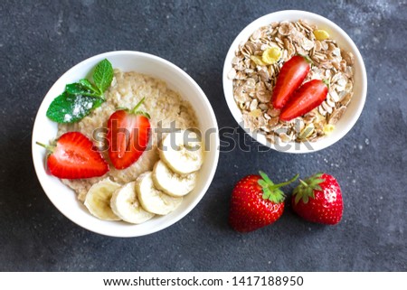 Oatmeal with strawberries and banana slices in a white plate standing on a dark concrete background. There is a plate with raw muesli, and strawberries are on the background.