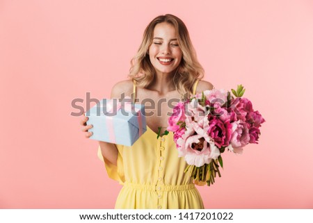 Image of a beautiful amazing young blonde woman posing isolated over pink wall background holding flowers holding present gift box.