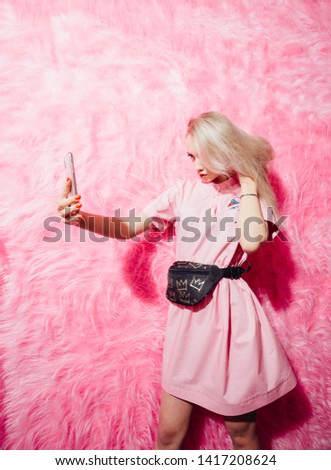 Beautiful young girl blogger dressed in fashion pink dress make a selfie on the background of pink fur wall in the show room