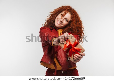 Portrait of alluring redhead woman 20s wearing leather jacket smiling and holding gift box isolated over white background