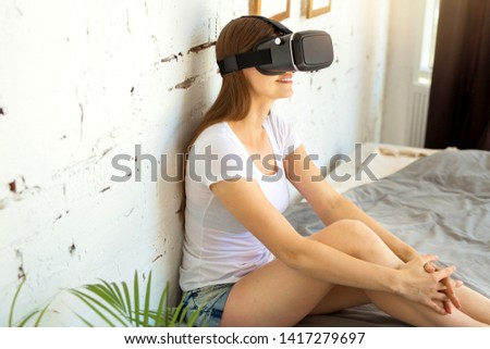 beautiful young woman in a white T-shirt sits on the bed with glasses of virtual reality indoors