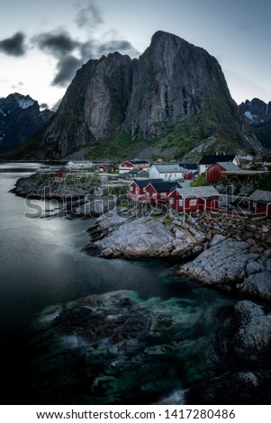 View on the beautiful fishermen village Hamnøy on Lofoten Islands in Norway with cute little red fisherman huts on pillars built on the shore of the ocean with a big mountain in the background