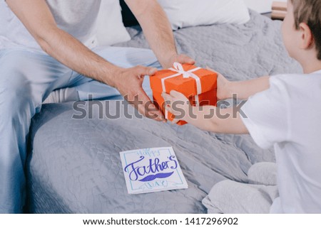 cropped shot of kid presenting fathers day gift box to daddy sitting on bedding