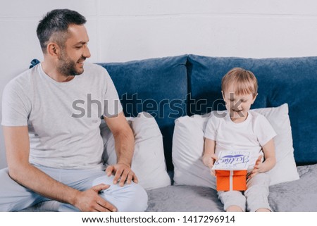adorable boy holding gift box and fathers day greeting card while sitting on bed near happy father