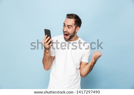 Image of a handsome excited young man using mobile phone isolated over blue wall background make winner gesture.