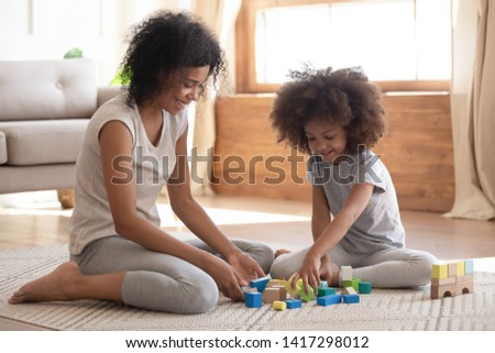 Cute little african american kid playing with black mom or baby sitter on floor, happy family mixed race mother helping small preschool child girl building constructor castle of wooden blocks at home