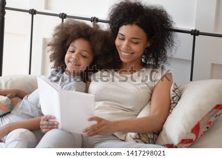 Happy african mother holding reading book relaxing in bed with cute child daughter, caring black parent mom tell small kid funny fairy tale bedtime story in bedroom having fun together in the morning