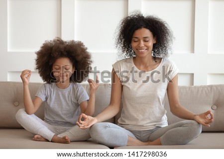 Healthy calm african american mother teaching meditation sit with cute small child daughter on sofa, mindful happy black mom and kid girl doing yoga exercises relaxing together in lotus pose at home Royalty-Free Stock Photo #1417298036