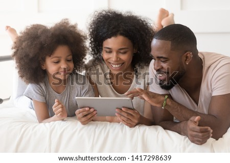 Happy african family with cute little kid daughter using digital tablet lying on bed together, black parents and small child girl having fun looking at computer screen in bedroom in the morning