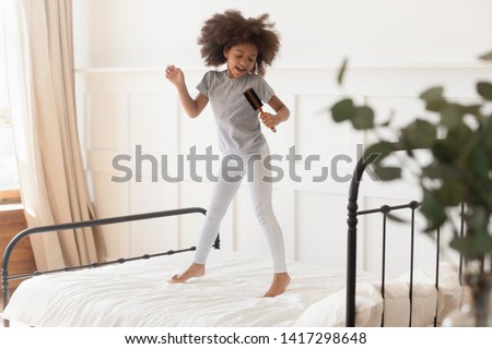 Happy funny cute little african american kid girl jumping on bed singing in microphone hairbrush at home, small mixed race child having fun dancing to music playing in bedroom alone in the morning