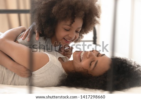 Happy cheerful mixed race family cute small kid daughter and young mum embracing playing lying on bed, funny african little child girl cuddling laughing hugging relaxing with joyful mother at home