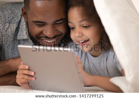 Happy family african father and cute small kid daughter using digital tablet lying on bed under blanket, funny little child girl with dad having fun looking at computer covered with duvet in bedroom