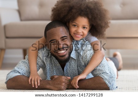 Cheerful african family single black father and cute funny little child daughter portrait, happy dad lying on carpet floor carrying small mixed race kid girl on back smiling looking at camera at home Royalty-Free Stock Photo #1417299128