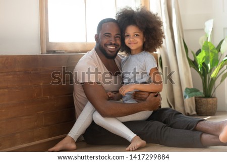 Happy african family young dad and cute little mixed race kid daughter bonding looking at camera sit on floor at home, small adorable child girl smiling on daddy lap, fathers day concept, portrait