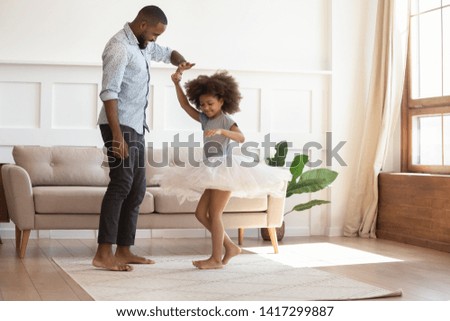 Loving black dad holding hand of child daughter dancing with his child princess wearing skirt, happy young african father and little cute daughter playing in living room enjoy time together at home Royalty-Free Stock Photo #1417299887