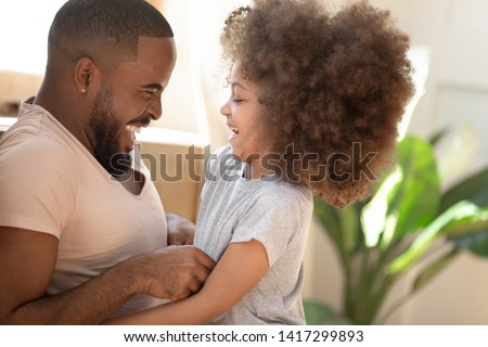 Cute small african american kid daughter laughing tickling black dad at home, cheerful little child girl having fun playing with happy daddy bonding enjoying funny moments on fathers day together