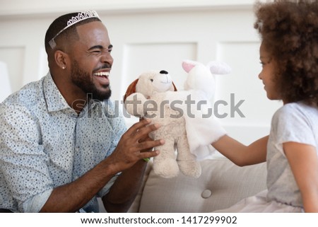 Happy funny african american father wearing crown holding toy having fun with cute small child daughter sit on sofa, cheerful black family dad and little kid girl laughing bonding playing at home