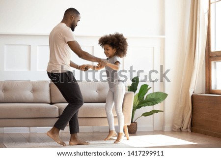 Happy african family father and cute little kid daughter jump having fun in living room in the morning, loving black dad holding hands of child girl dancing together enjoy funny activity play at home