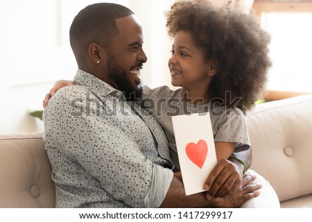 Happy african family dad and little cute kid daughter laughing embracing celebrate fathers day together, cheerful young black dad holding greeting card with red heart congratulation from small child