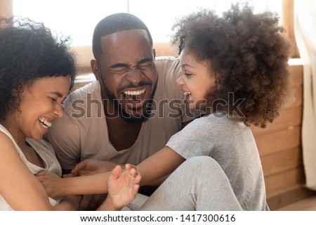 Happy african american family with cute little kid daughter tickling laughing at home together, cheerful black parents and small child girl having fun playing bonding enjoy funny lifestyle activity Royalty-Free Stock Photo #1417300616