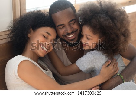 Happy affectionate african american family of three bonding embracing, cute kid child daughter and loving parents cuddling congratulating dad with fathers day hugging smiling daddy laughing together Royalty-Free Stock Photo #1417300625