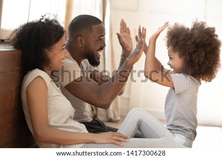 Happy african american family mom dad and little cute kid girl having fun at home, mixed race small child daughter give high five to daddy play with parents laugh enjoy warm moments in the morning