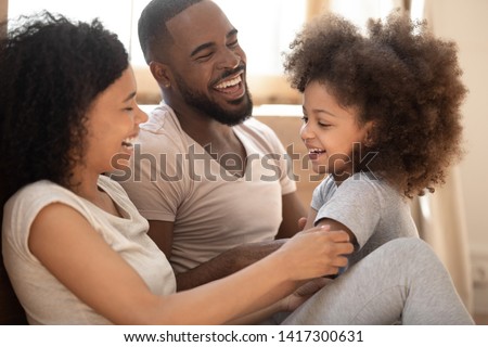 Loving black family relaxing bonding in bedroom in the morning, happy african american parents laughing cuddling having fun with cute little kid child daughter playing enjoying moments together