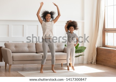 Carefree funny african family young mom having fun with cute little kid girl jump dancing in living room, happy mixed race mother with small child daughter laugh do morning exercise together at home Royalty-Free Stock Photo #1417300847