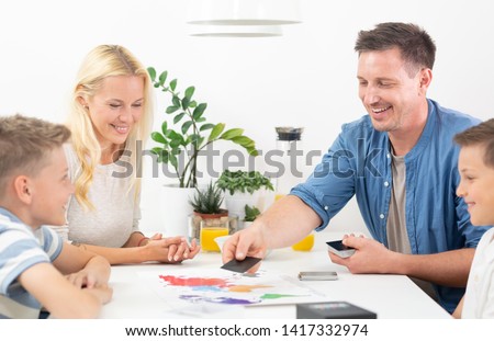 Happy young family playing card game at dining table at bright modern home. Spending quality leisure time with children and family concept. Cards are generic and debranded. Royalty-Free Stock Photo #1417332974