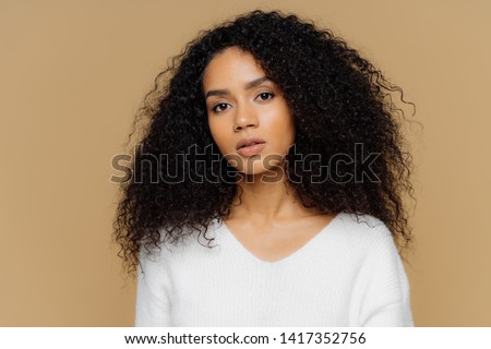 Portrait of serious beautiful dark skinned female with frizzy black hair, has minimal makeup, looks calmly at camera, wears white jumper, stands against brown background, being deep in thoughts. Royalty-Free Stock Photo #1417352756
