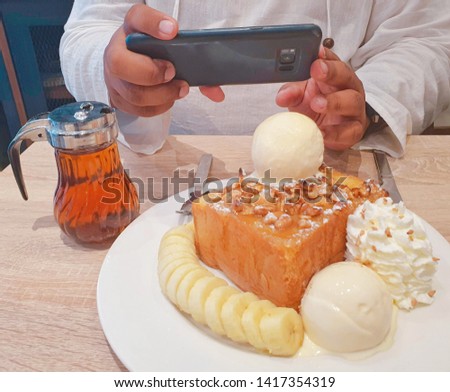 Man using smartphones to take pictures of honey toasts with bananas and ice cream at modern wooden tables in coffee shops.