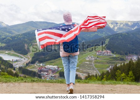 Outdoor shot of slim girl at nature background with American flag on her shoulders, woman wearing casual rosy jacket, trousers, sneackers and rucksack having vocation in mountains. Traveling concept.