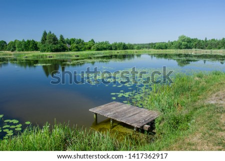 Wooden small bridge with planks at the pond, trees on the horizon and clear blue sky