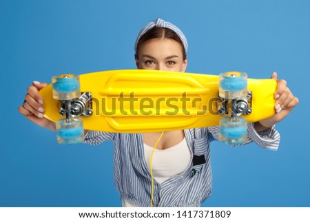 Photo of beautiful young woman hold yellow penny or skateboard in front of face over blue background.