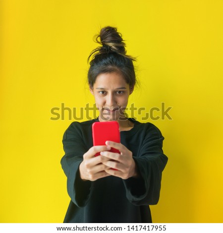 Cheerful young brunette girl with mobile phone, smiling, taking selfie, posing on yellow background