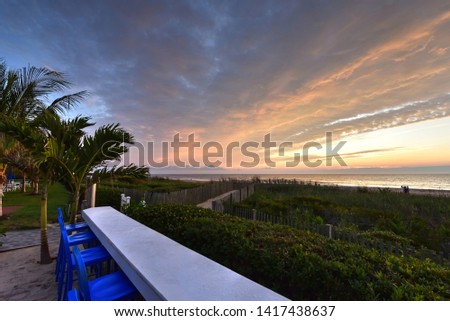 An empty beach bar views colorful sunrise skies on the Atlantic Ocean. Blue bar stools contrast a white counter. Palm trees frame the left. Hues of orange, yellow & blue color the scenic morning sky.